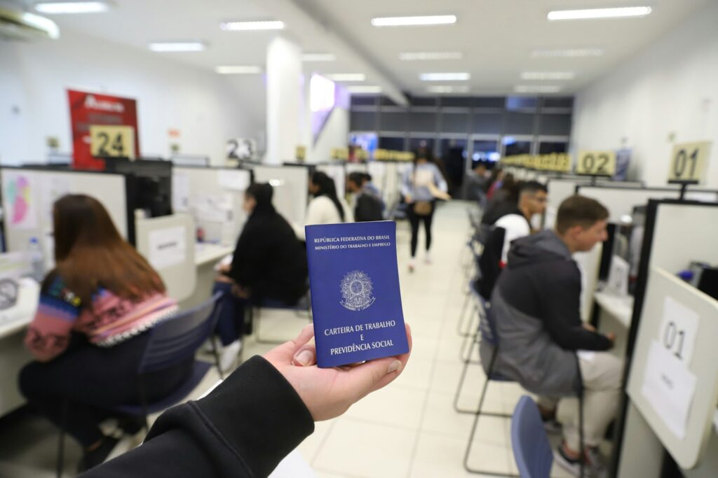 Em primeiro plano, uma mão de pele branca segura uma Carteira de Trabalho (CLT). Ao fundo, desfocadas, pessoas são atendidas em guichês.